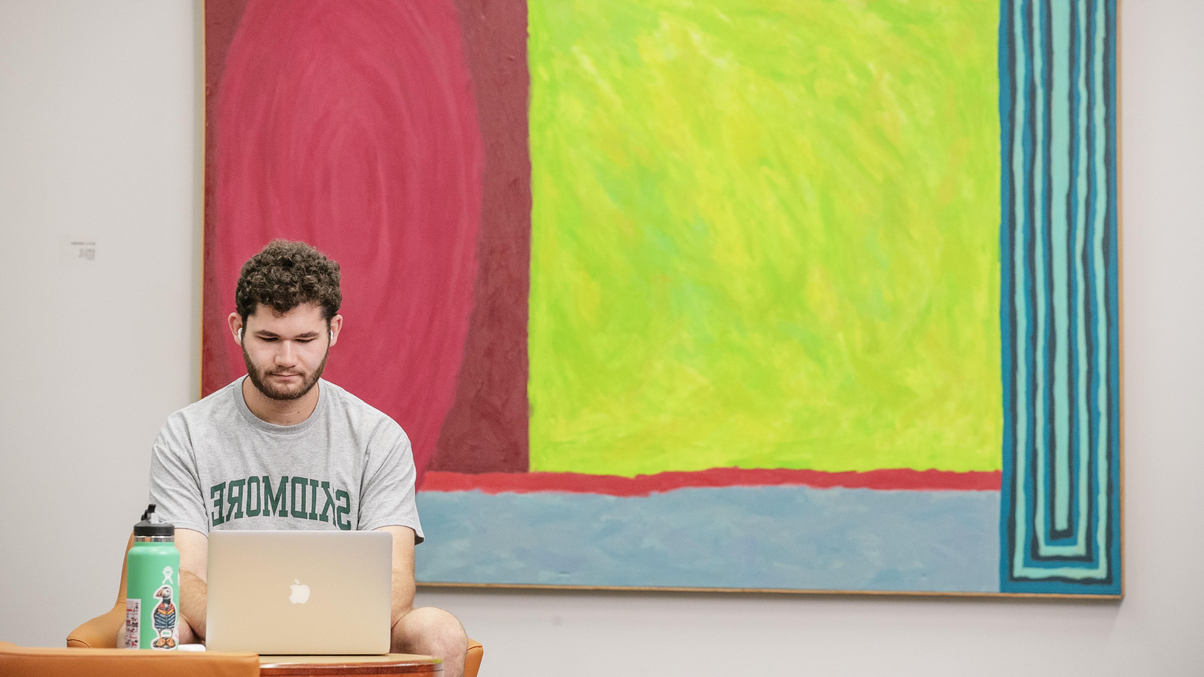 Milo Flamenbaum ’26 sits beside a large abstract painting in Lucy Scribner Library, working on a laptop.